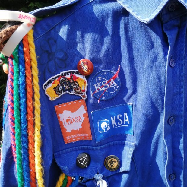 Schildjes Het blauwe schildje van KSA Nationaal en het oranje schildje van KSA Oost-Vlaanderen komen boven het rechterborstzakje.